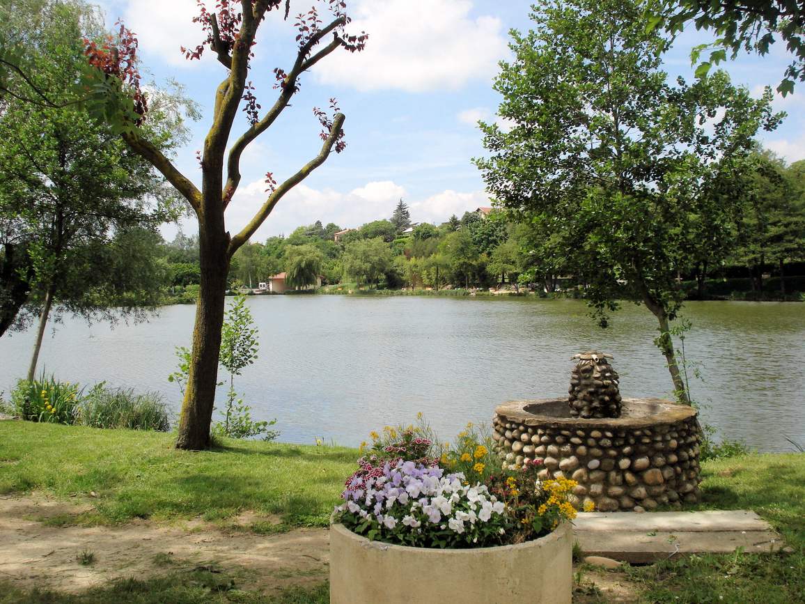 L'étang d'Anneyron bordé d'arbres, avec une fontaine en galets au premier plan